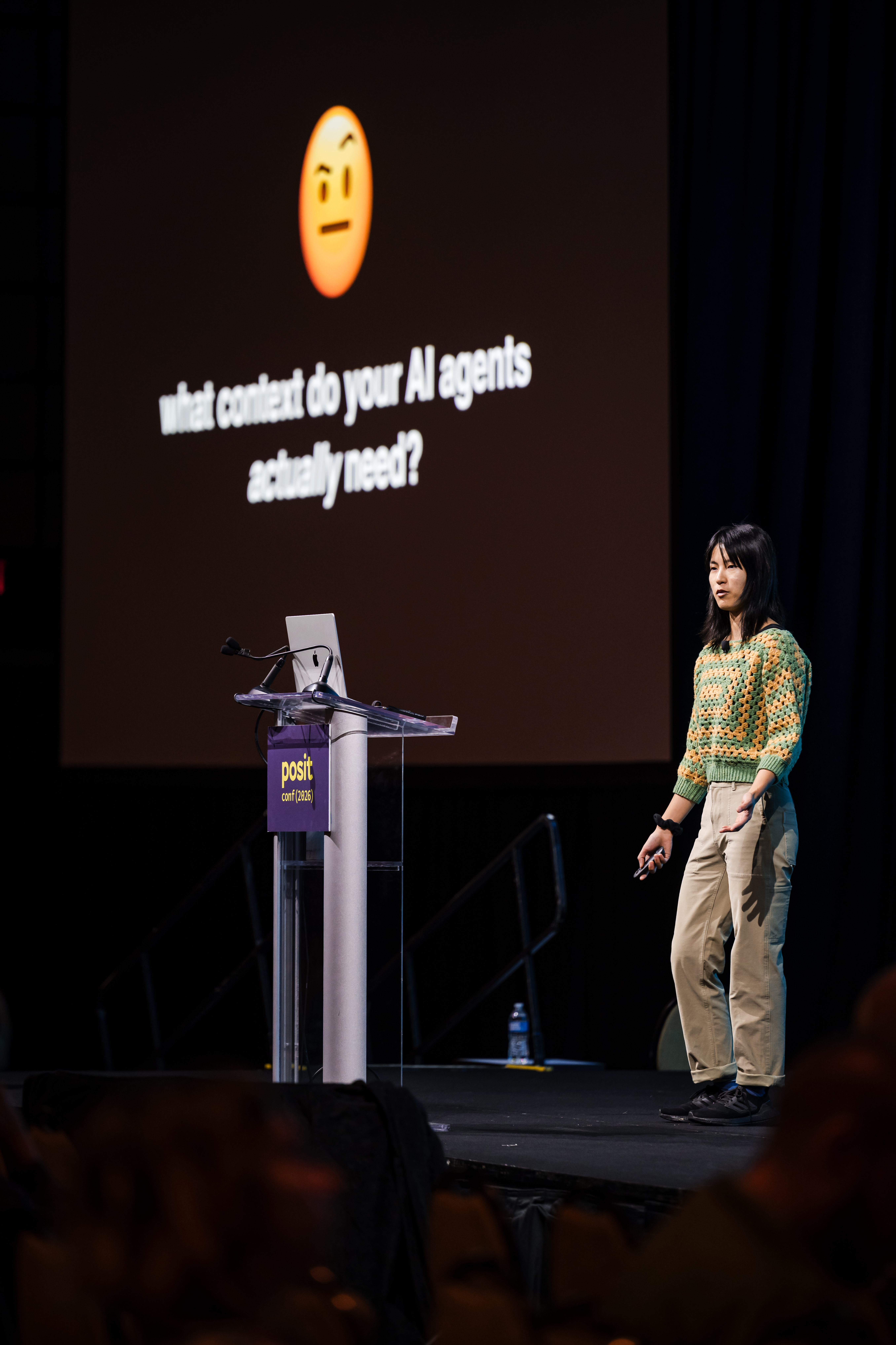 A speaker standing on a stage next to a Posit-branded podium gives a presentation with a backdrop reading "what context do your AI agents actually need?"