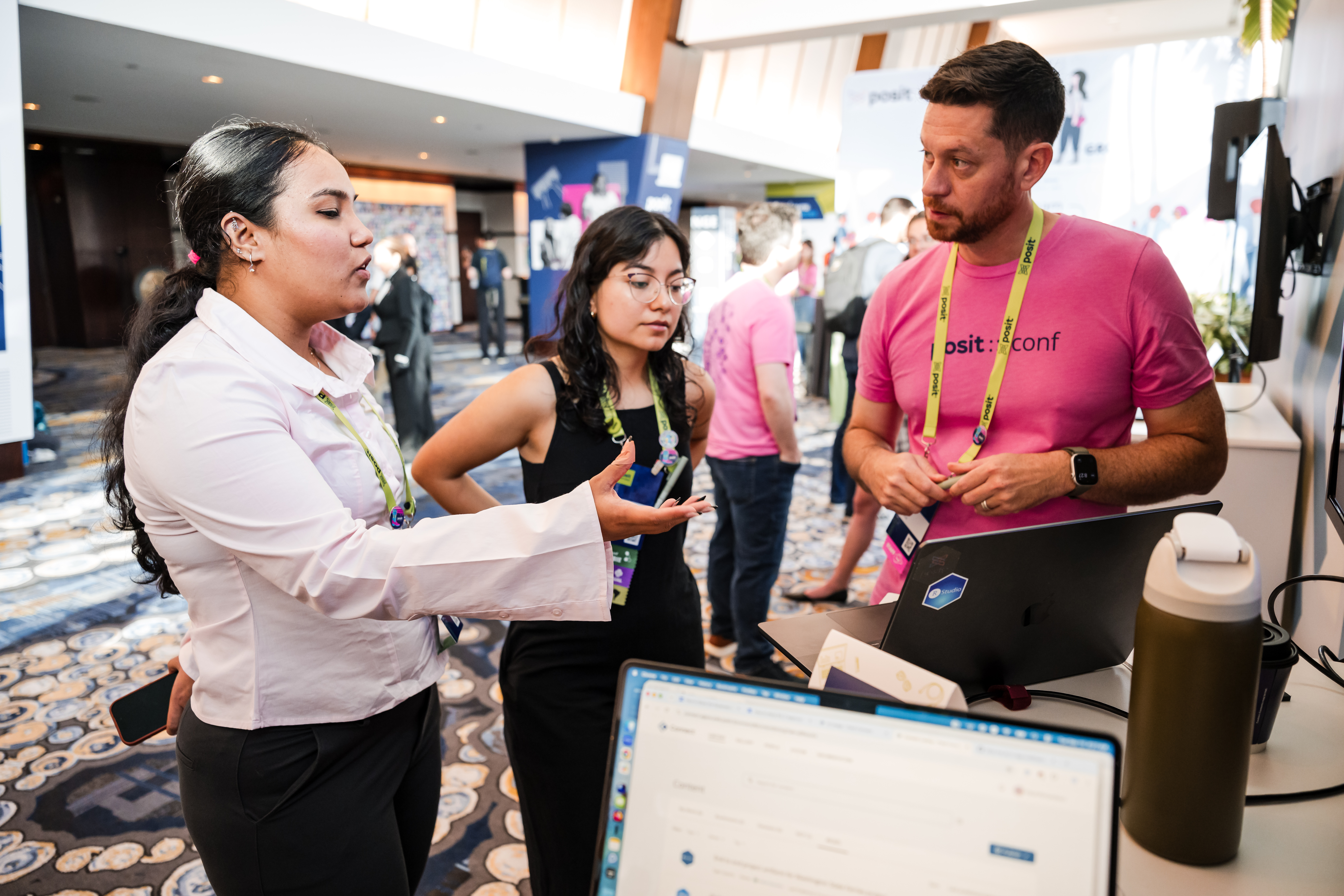 A person in a white shirt gestures while talking to a person in a pink posit::conf shirt at a conference booth, with another attendee listening between them.