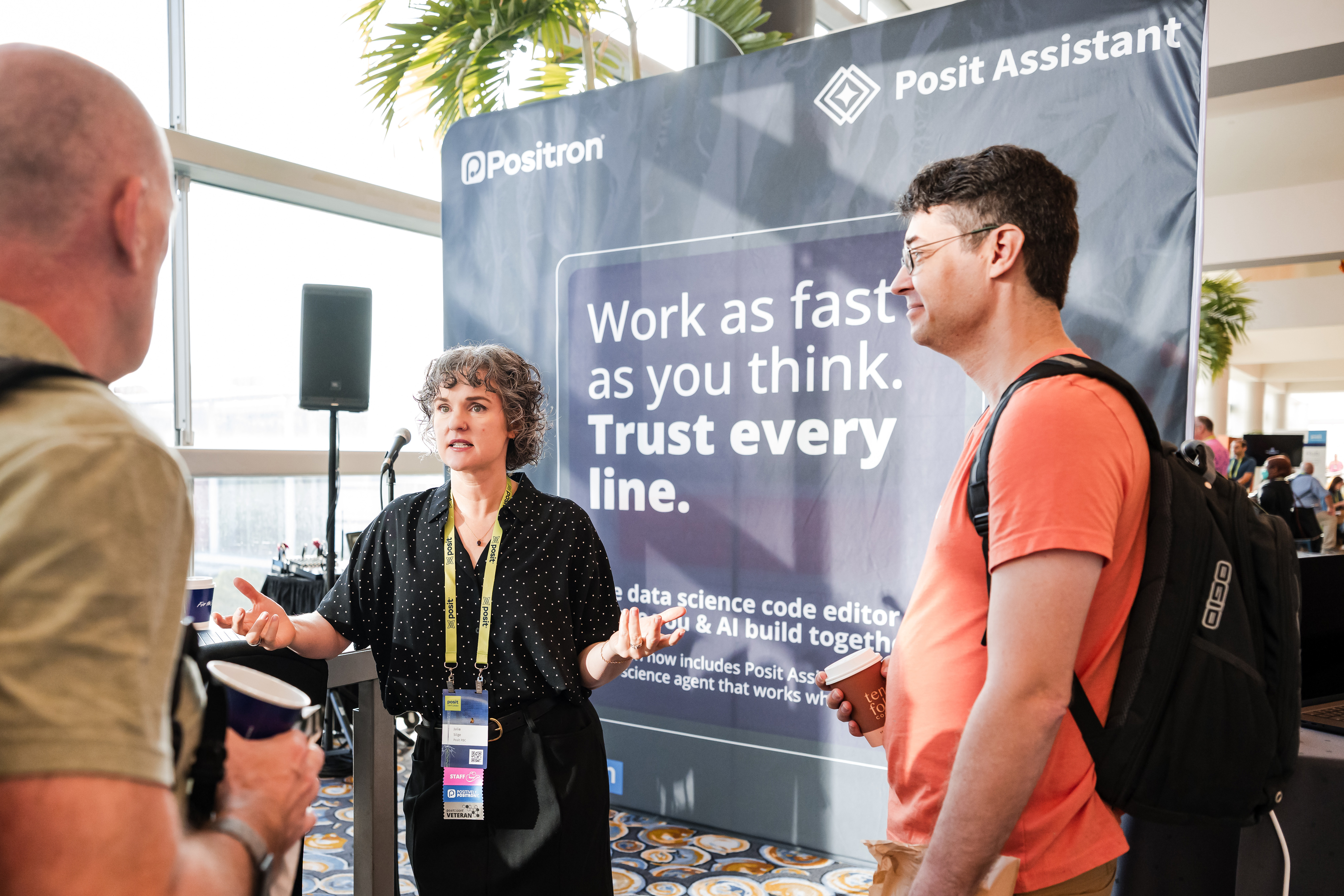 A woman wearing a conference lanyard speaks with two attendees at a Positron booth, with a backdrop reading 'Work as fast as you think. Trust every line.'