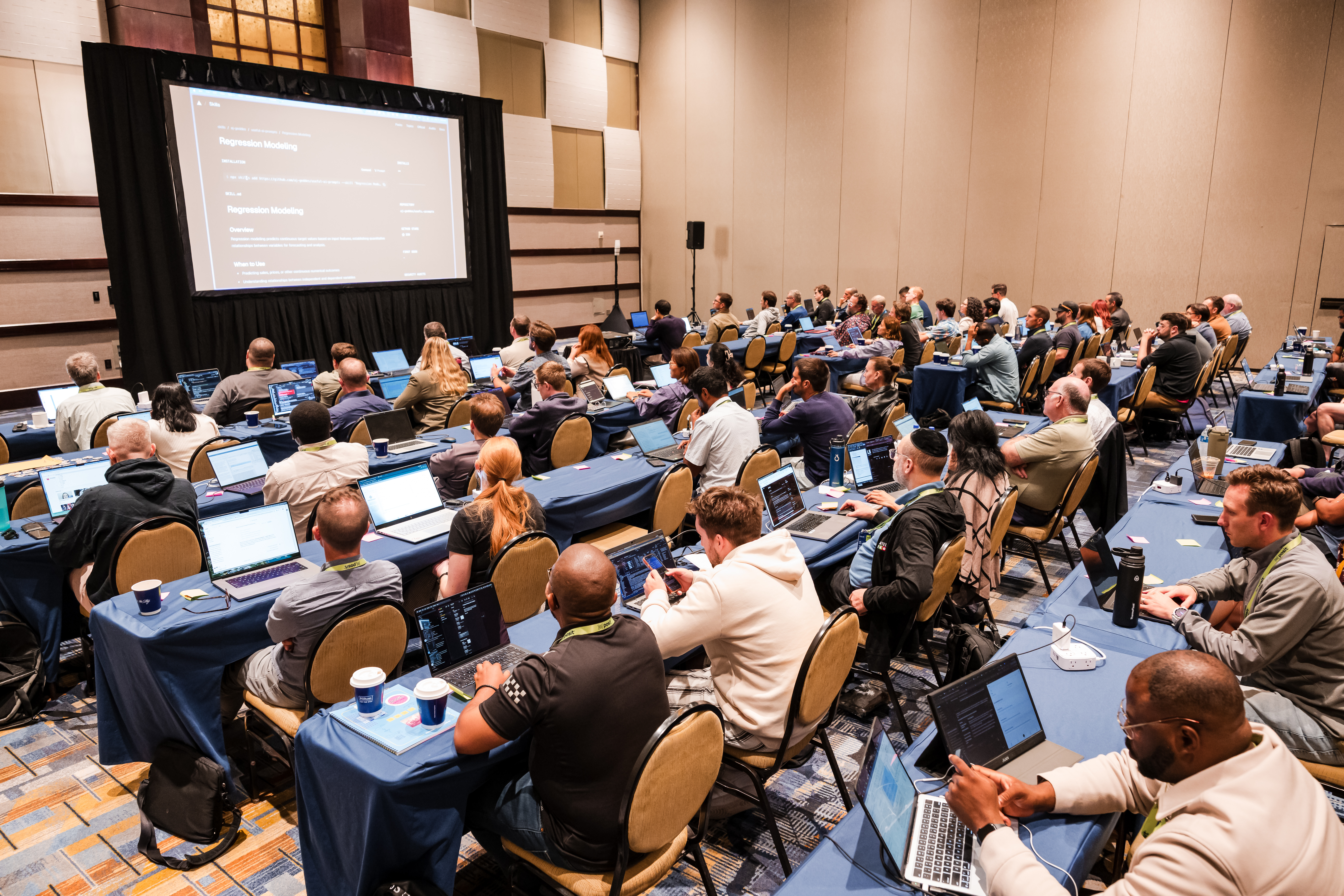 High-angle view of a full conference workshop room with rows of attendees sitting at long, cloth-covered tables with open laptops. Most participants are working or looking toward a large projection screen at the front of the room displaying presentation slides and code.