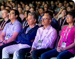 A diverse audience of professionals sitting in a darkened auditorium at a conference. The foreground shows four men in a row, including one in a vibrant pink shirt, all looking toward a stage with focused expressions. They are wearing conference lanyards and badges. The background is filled with a large, blurred crowd of attendees, creating a sense of a major industry event.