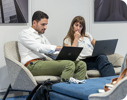 Two professionals sitting in comfortable lounge chairs, focused on their respective Dell laptops. The man on the left points toward his screen while speaking to the woman on the right, who looks on thoughtfully. They appear to be in a modern office or conference setting with clean white walls and framed architectural photography in the background.