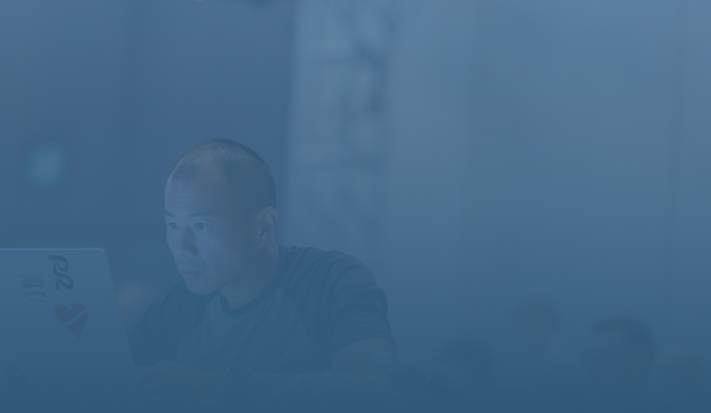 A blue-tinted, monochromatic image of a man focused on his laptop in a workspace.