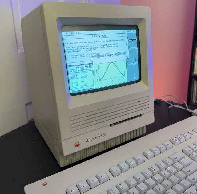 A beige Macintosh SE/30 computer with a connected keyboard. The screen shows a graphical interface with several windows open, including one titled "Mac of Beyond" and another with a graph. The time "4:23 PM" is visible in the menu bar.