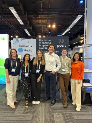 A group of six smiling professionals posing together at a conference booth. They are standing in front of a backdrop that reads "The future of pharma is open source" with the Posit and RStudio logos. The group is dressed in business casual attire and wearing conference lanyards. The setting is an indoor event space with industrial ceilings and grey carpeting.
