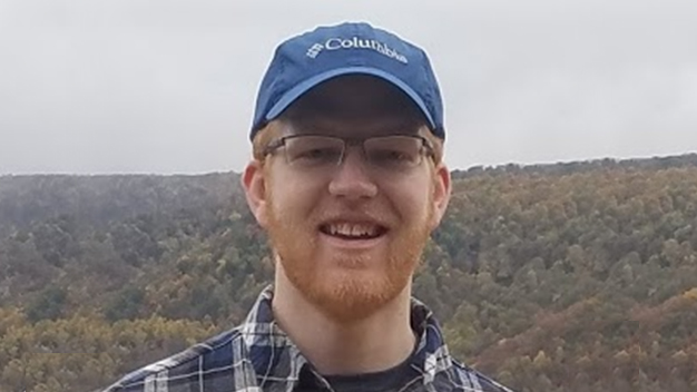 portrait of Nate Kratzer standing outside in front of a hill covered in trees