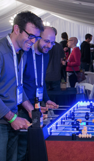 Two men with event badges smiling and playing foosball at a conf event