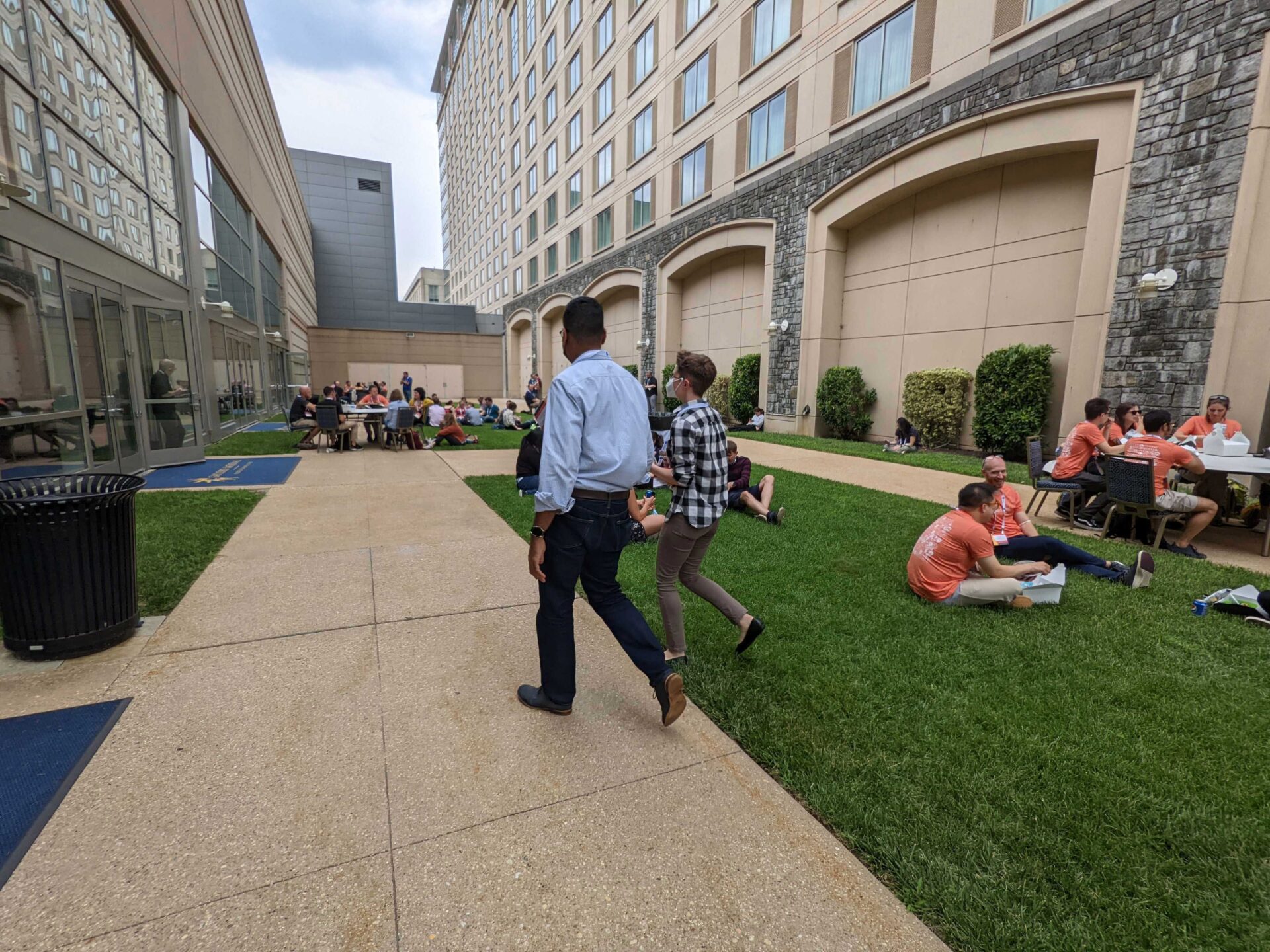 2 people walking while others sit at a table and on the grass outside in between 2 buildings