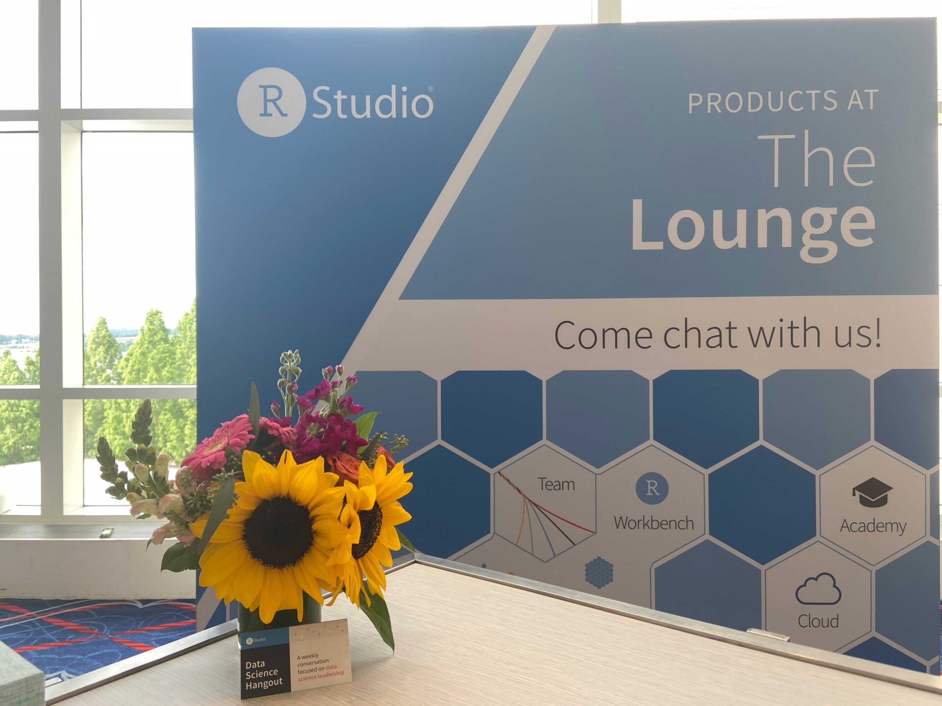 Text: RStudio Products at The Lounge, Come chat with us! Team, Workbench, Academy, Cloud, booth backdrop behind table with flowers and Data Science Hangout card