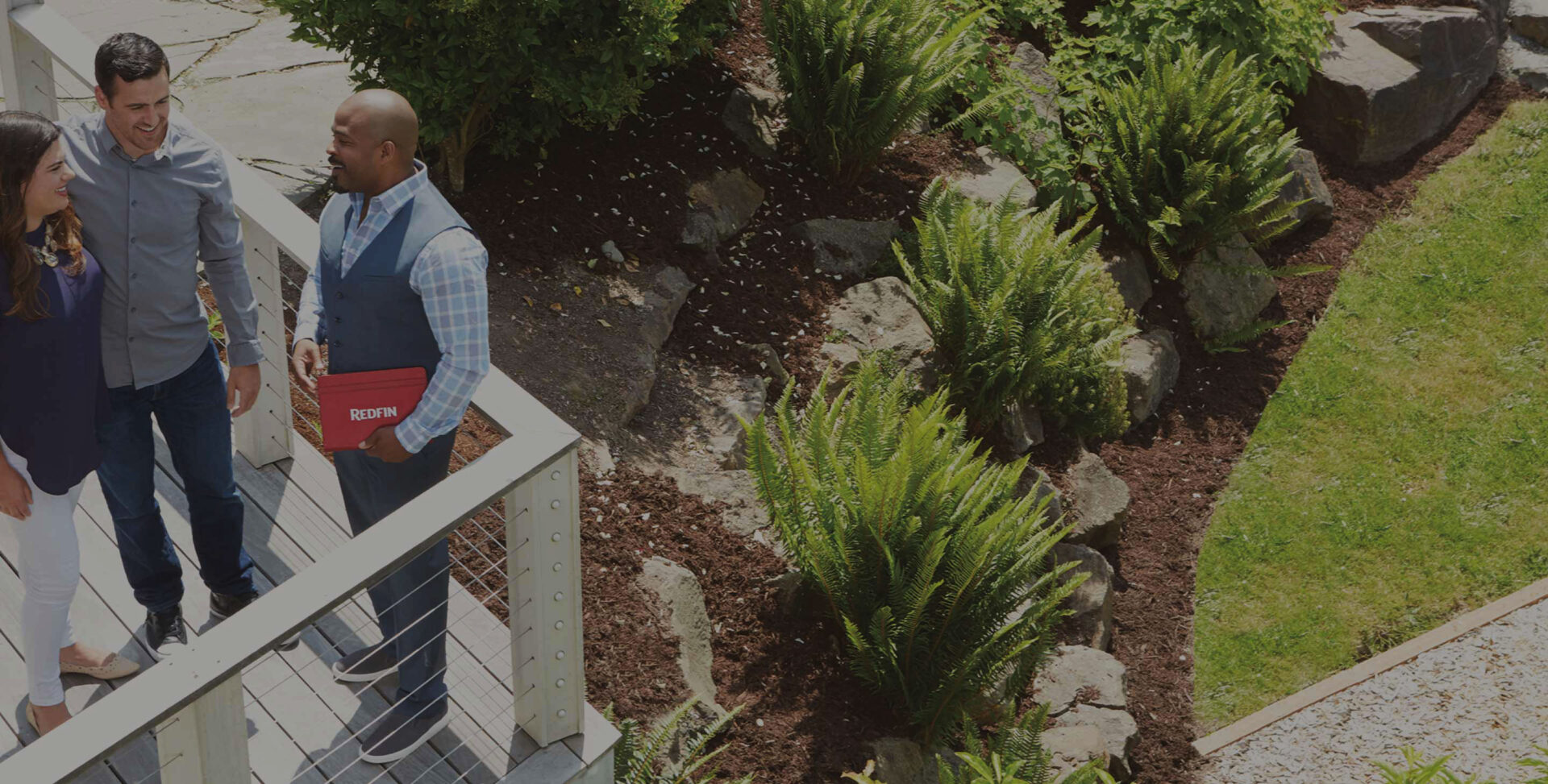 photo of realtor holding Redfin folder talking to couple on a balcony above a landscaped yard