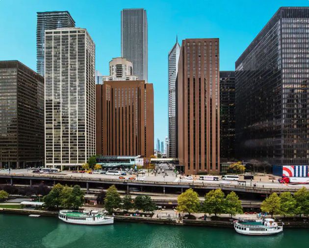 Semi-aerial angle of Downtown Chicago waterfront with large hotels and office buildings side by side in front of a waterway with two ferries.